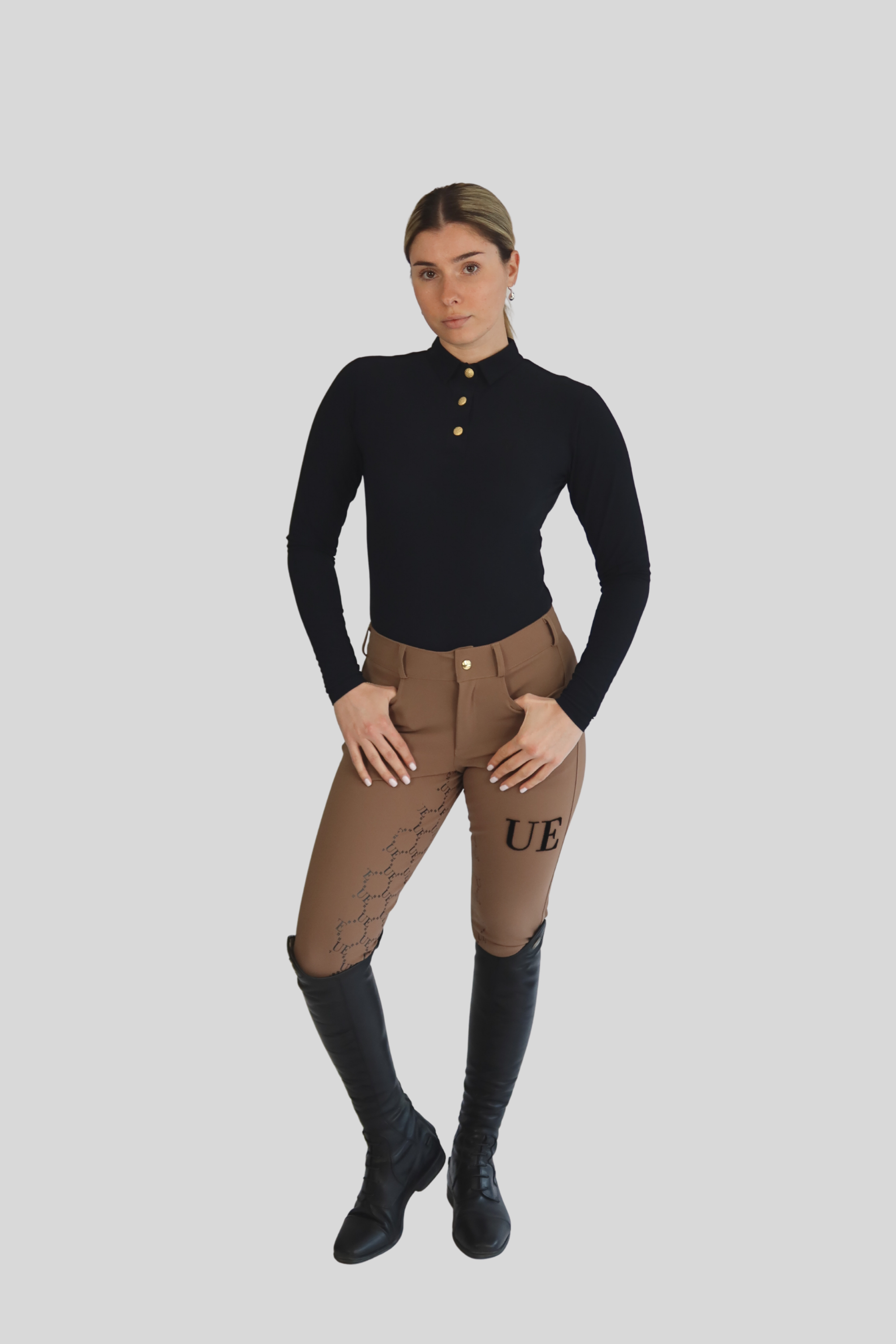 Person wearing a black top and brown riding pants with 'UE' branding on a gray background